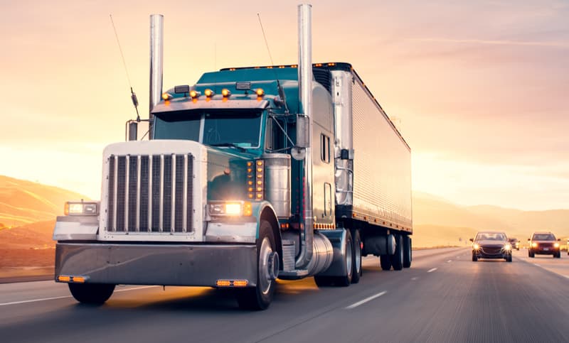 Large semi‑truck driving along a highway at sunset, with cars following behind and hills in the background.