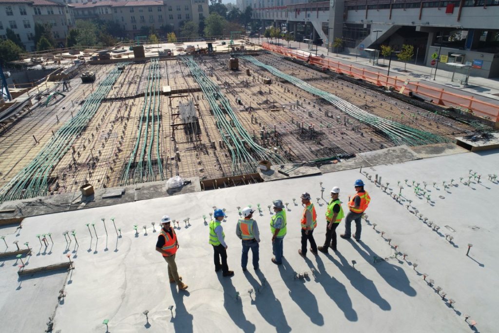 Group of construction workers wearing safety vests and helmets standing on a concrete slab, inspecting progress at a large building site with rebar and conduit laid out