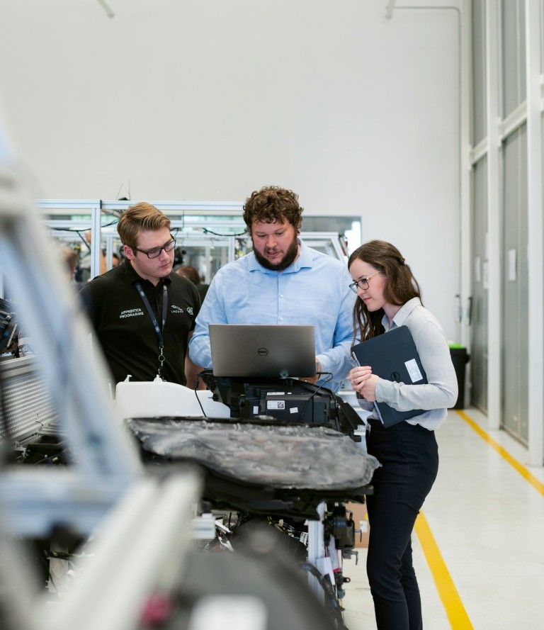 Three engineers collaborating around a laptop in a bright industrial workspace, discussing technical data beside machinery.