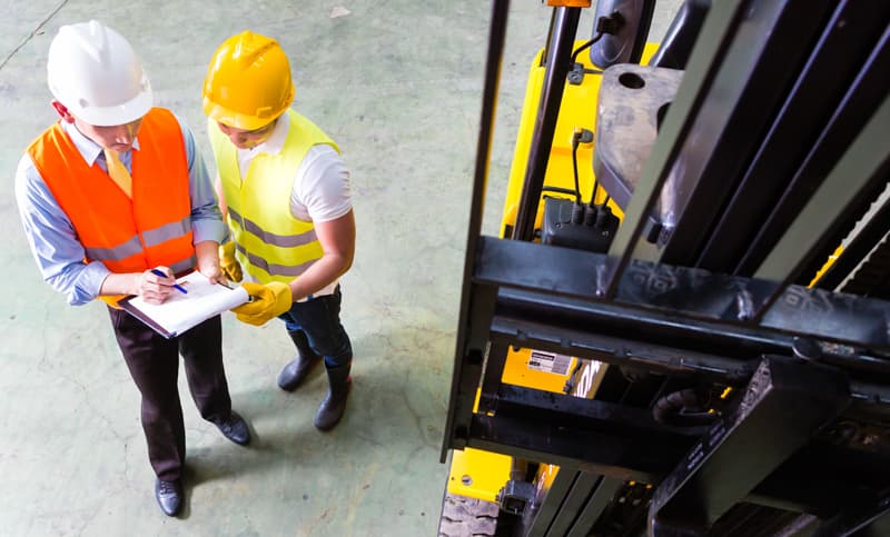 Two workers wearing hard hats and reflective vests reviewing notes on a clipboard beside a yellow forklift in an industrial warehouse.