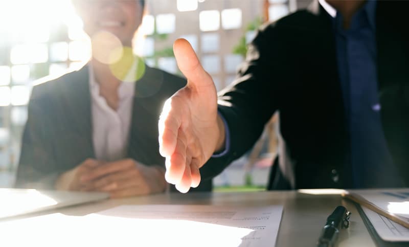 Businessperson extending a hand for a handshake across a desk with documents and a pen, symbolizing agreement or partnership in a bright office setting.