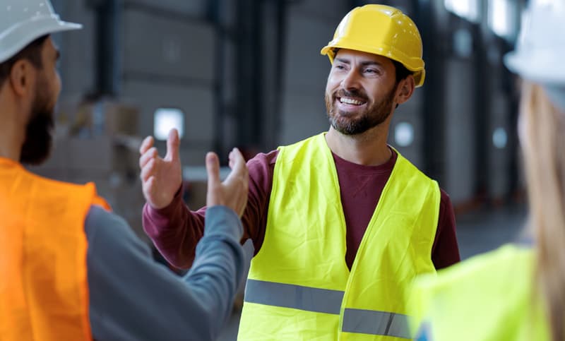 Smiling construction worker wearing a yellow hard hat and reflective vest greeting colleagues inside an industrial warehouse.