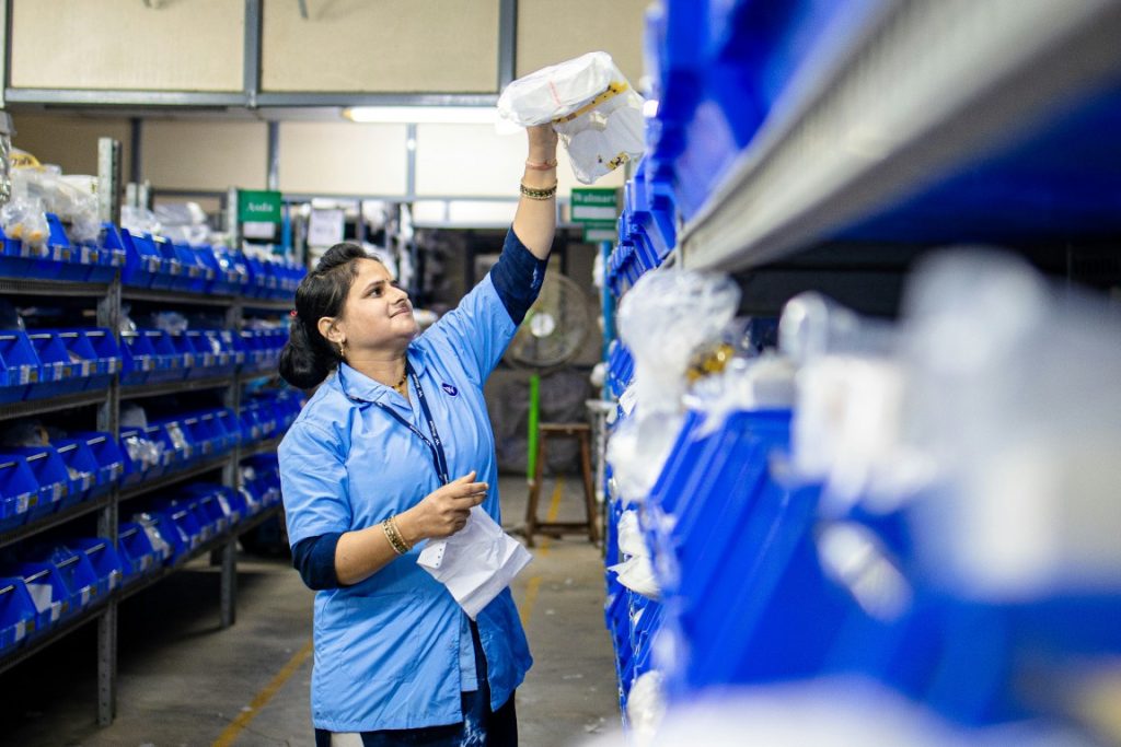 Warehouse worker wearing a blue uniform reaching for an item on a high shelf filled with blue storage bins, checking inventory with a paper list.