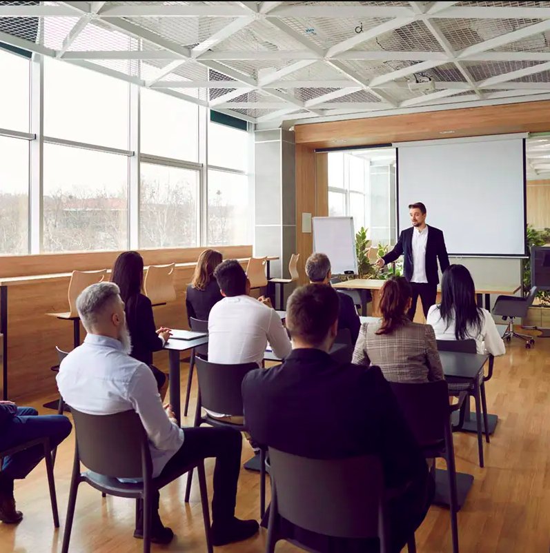 Business professional giving a presentation to a group of people seated in a conference room with large windows and a projection screen.